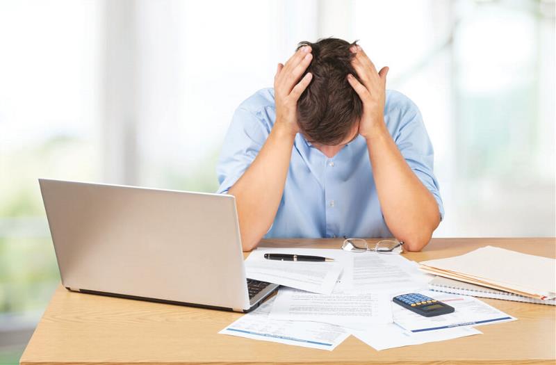 A frustrated man holding his head with his laptop, calculator and sheets of paper scattered on his table