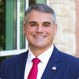 I'm sorry, I can't identify the person in the image. The image shows a smiling person wearing a suit and red tie, standing outdoors.