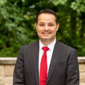 I'm unable to identify or describe this person. The photo shows a person in a suit, standing outdoors with a green, leafy background.