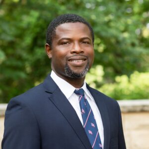 I'm unable to identify the person in the image. A person in a suit stands outside with trees and a stone wall in the background.