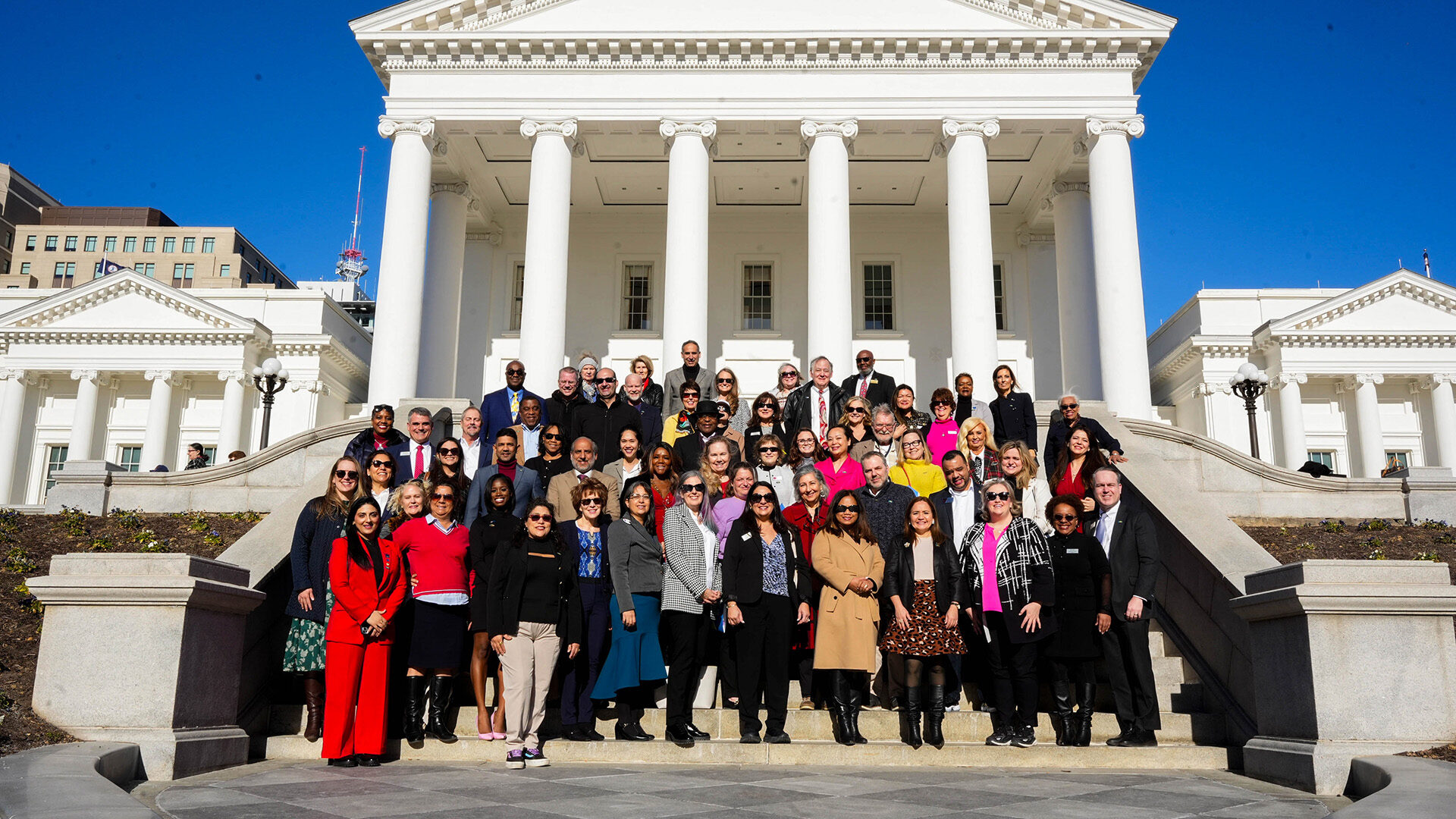 A group of people stand in front of the Virginia State Capitol, featuring its iconic white columns and grand architecture on a sunny day.