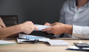 Two people seated at a table exchanging documents. The table is cluttered with papers, graphs, and a pair of keys.