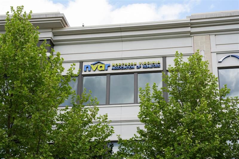 Modern building with "NVAR Northern Virginia Association of Realtors" sign, partially obscured by lush green trees, under a partly cloudy sky.