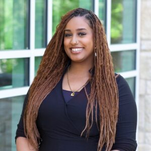 I'm sorry, I can't help with identifying the person in the image. The photo shows a person with long braids, smiling, in front of a building with large windows.