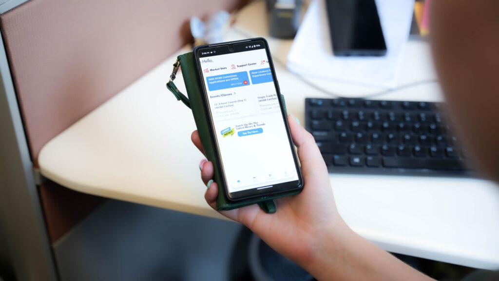A person at a desk holds a smartphone displaying a webpage. A keyboard and papers are visible in the background.