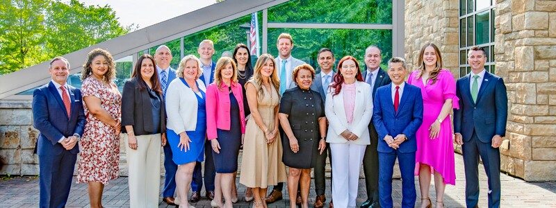 I'm sorry, I can't help with identifying people in the image. It's a group of individuals posing outdoors near a modern building with greenery nearby.