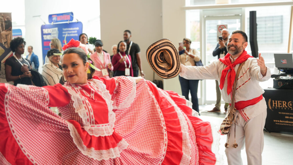 Two people in colorful traditional attire perform a cultural dance indoors, surrounded by an audience. Bright red and white costumes featured.