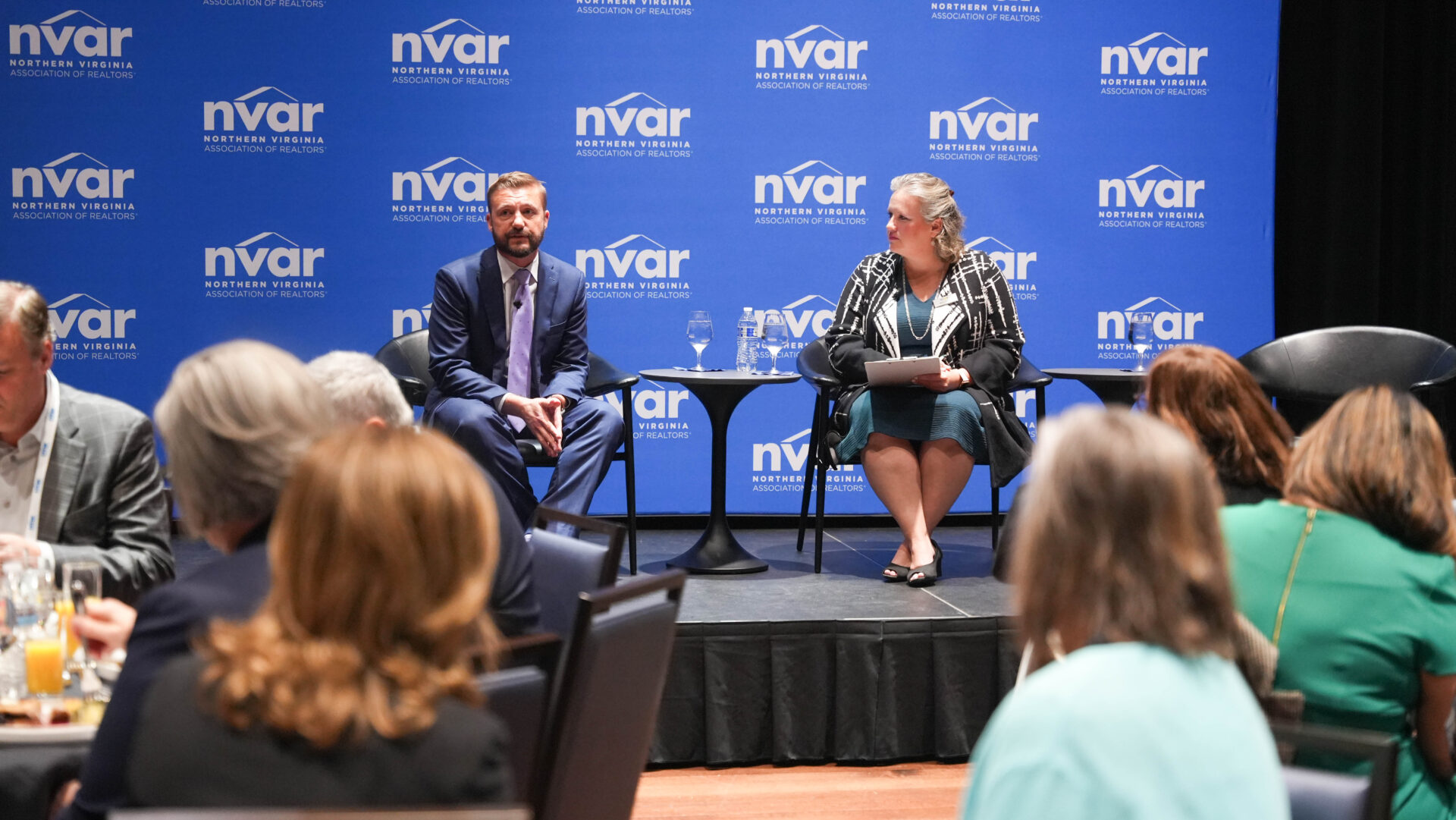 Two people are seated on stage during an NVAR event, engaging with an audience in a conference setting.