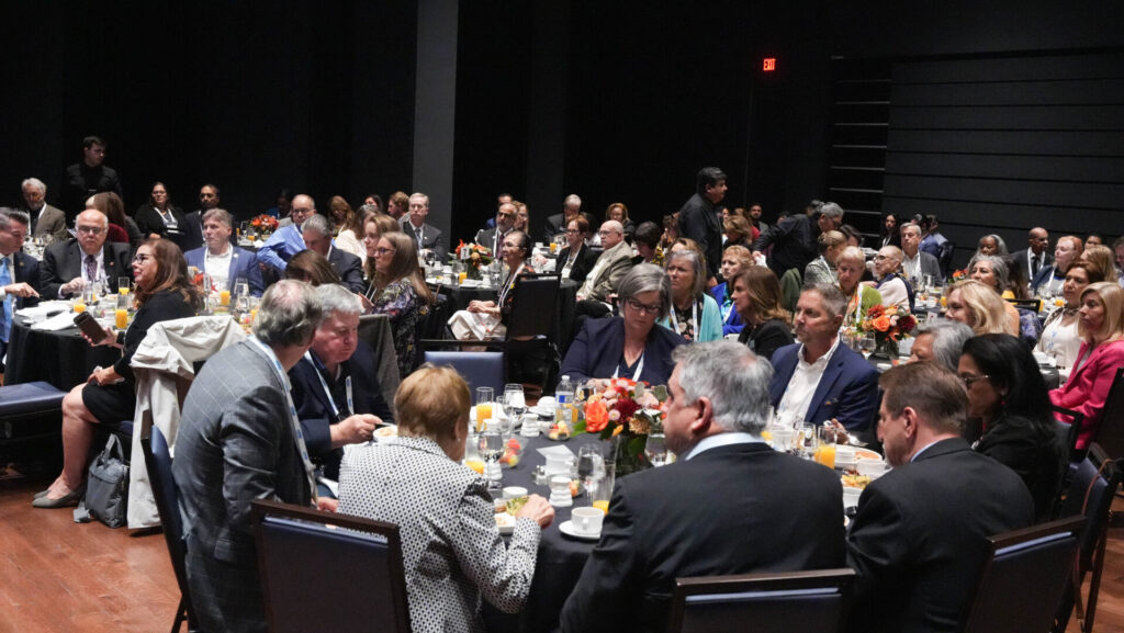 A large group of people seated at tables, engaged in a formal gathering or conference, with a dark background and floral table arrangements.