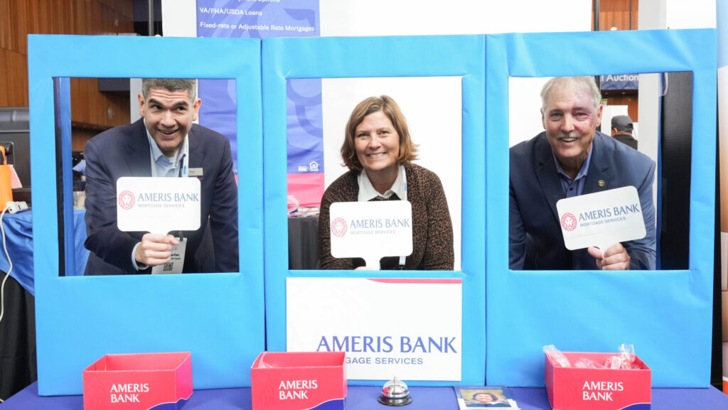 Three people smiling through blue frames, each holding an Ameris Bank sign, with bank promotional items displayed in front of them.