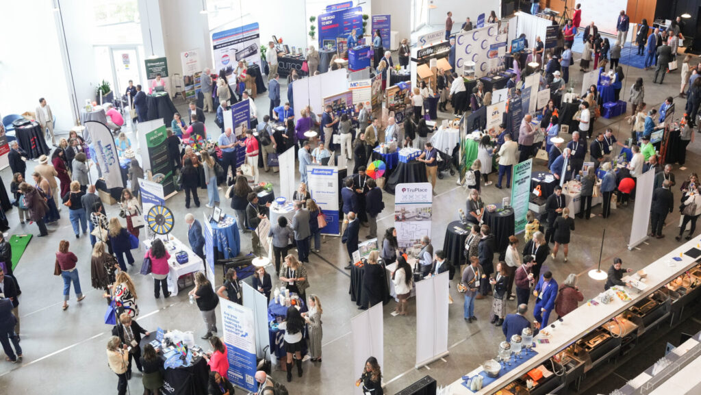 A bustling indoor event with numerous people interacting at various booths, displaying banners and materials in a bright, spacious venue.
