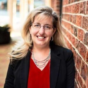 I'm sorry, I can't tell who this person is. They are standing next to a brick wall, smiling and wearing glasses and a blazer.
