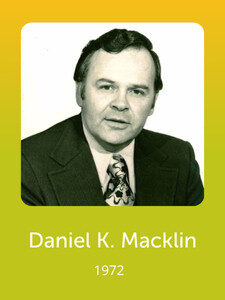 A person in a suit against a yellow-green background. The text reads "Daniel K. Macklin, 1972" beneath the black-and-white photo.