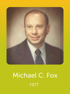 A person in a formal suit poses for a vintage-style photograph. The background is plain. Text below reads "Michael C. Fox, 1977."