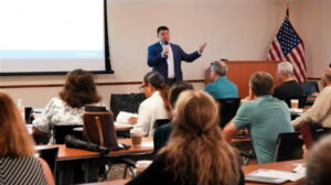 A person speaks in front of a classroom. An audience listens attentively. An American flag is visible in the background.