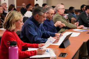 A group of people seated at tables, engaged in a meeting, taking notes on documents. A pink water bottle and tablet are visible.