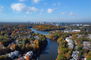 Aerial view of a winding river surrounded by colorful autumn trees, suburban homes, and a distant city skyline under a clear blue sky.