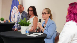Four people seated at a panel discussion, one holding a microphone, with Virginia state flag in the background and small plants on tables.
