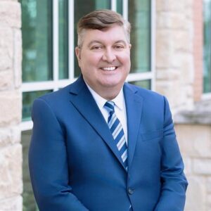 A person in a blue suit stands smiling outdoors, against a brick and stone building background with windows reflecting light.