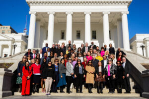 A group of people stands on the steps of the Virginia State Capitol, a neoclassical building with columns, under a clear blue sky.