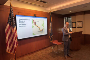 A person stands near a presentation screen and flags in a conference room, discussing "Delegate Marcus Simon's Road to Richmond Luncheon."