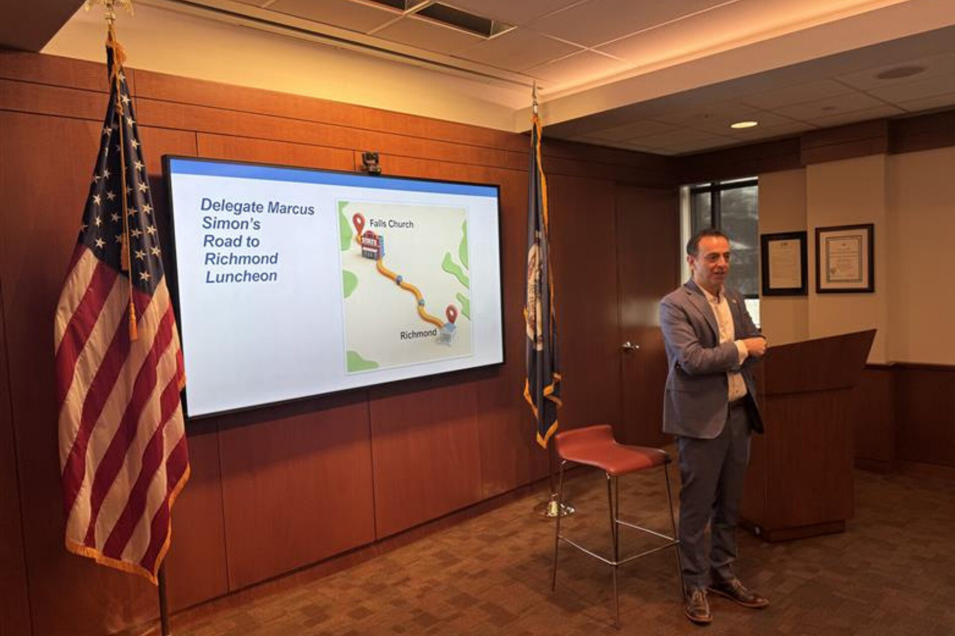 A person stands near a presentation screen and flags in a conference room, discussing "Delegate Marcus Simon's Road to Richmond Luncheon."