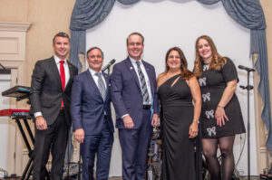 Five people in formal attire stand together in a room with musical instruments in the background. They appear to be at an event.