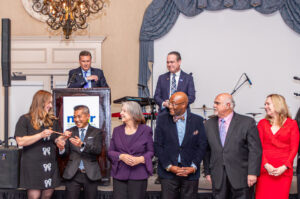 Seven people stand on a stage, smiling, with one speaking at a podium. A chandelier hangs above in an elegantly decorated room.