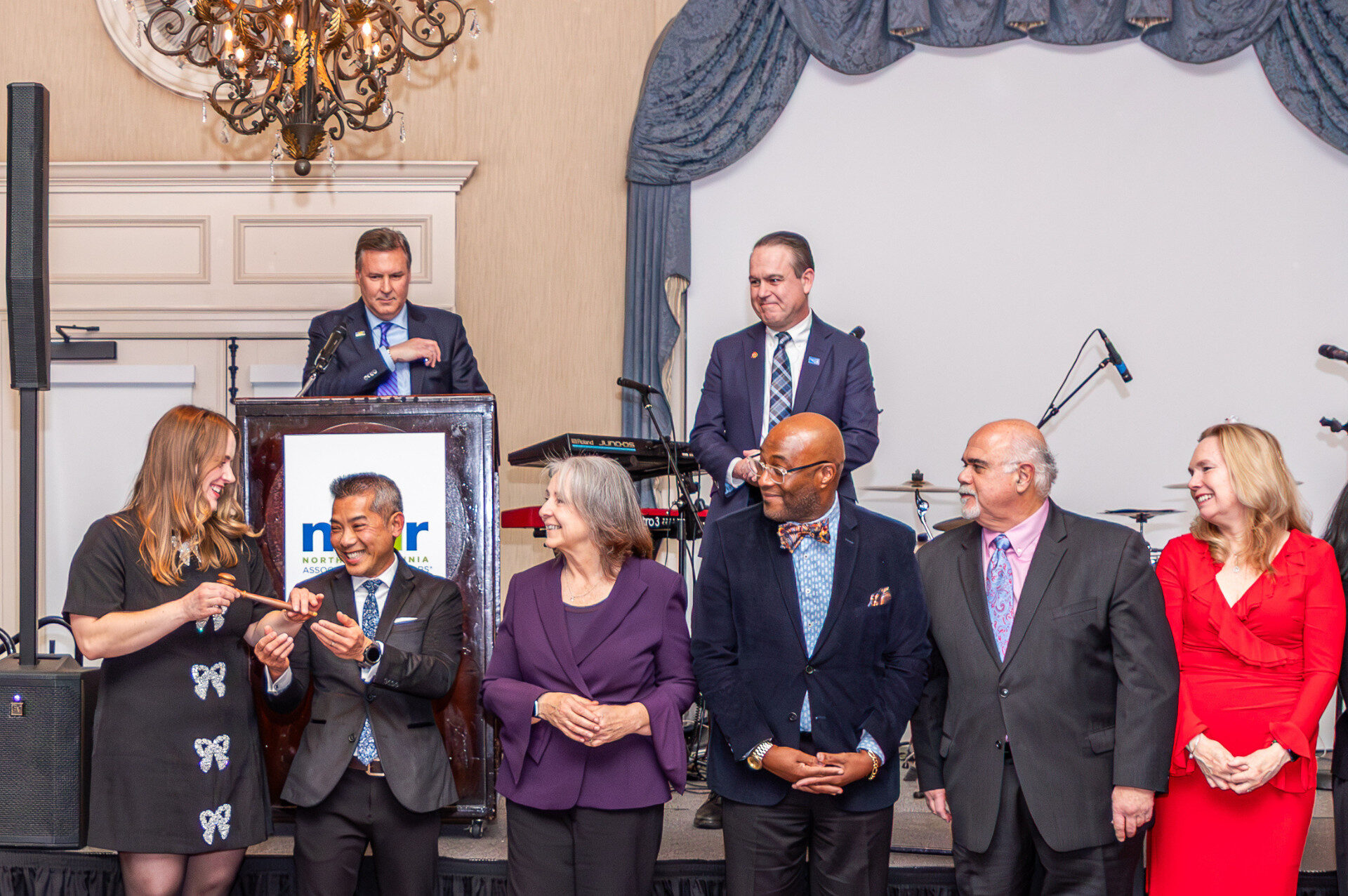 Seven people stand on a stage, smiling, with one speaking at a podium. A chandelier hangs above in an elegantly decorated room.
