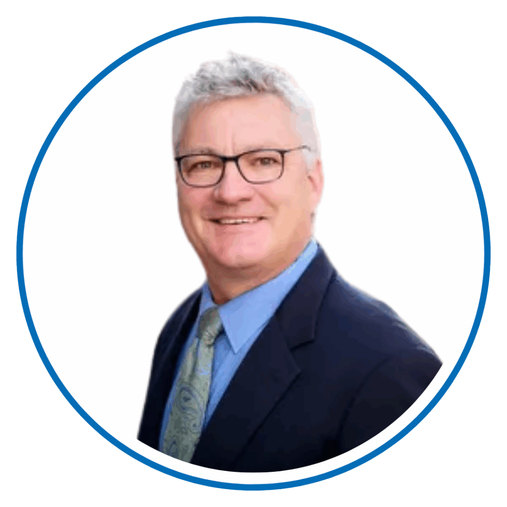 A person with gray hair, glasses, and a suit smiles against a white background. The image has a circular border with a blue outline.