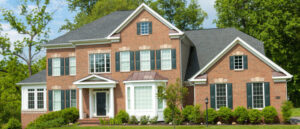 A large, two-story brick house with black shutters and white trim, surrounded by green trees and shrubs, sits on a well-manicured lawn.