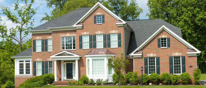 A large, two-story brick house with black shutters and white trim, surrounded by green trees and shrubs, sits on a well-manicured lawn.