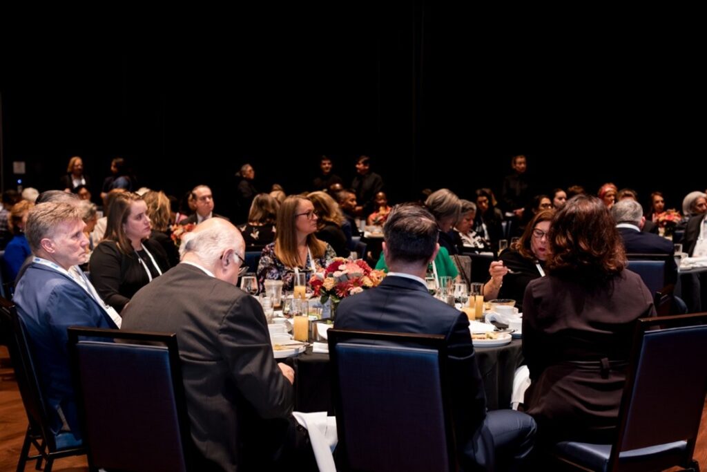 People seated at round tables during an indoor event, with flowers and table settings. Attendees are engaged, suggesting a formal or business gathering.