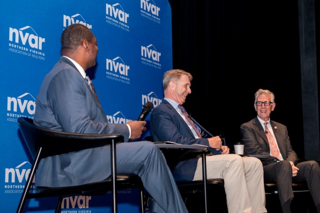 Three people sit and converse at a Northern Virginia Association of Realtors event with blue-logoed backdrop and microphones, engaging in a panel discussion.