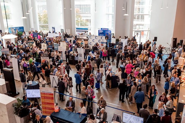 A bustling indoor event with many people interacting, booths displaying information, tall windows, and natural light. Modern architecture, no recognizable landmarks or buildings.