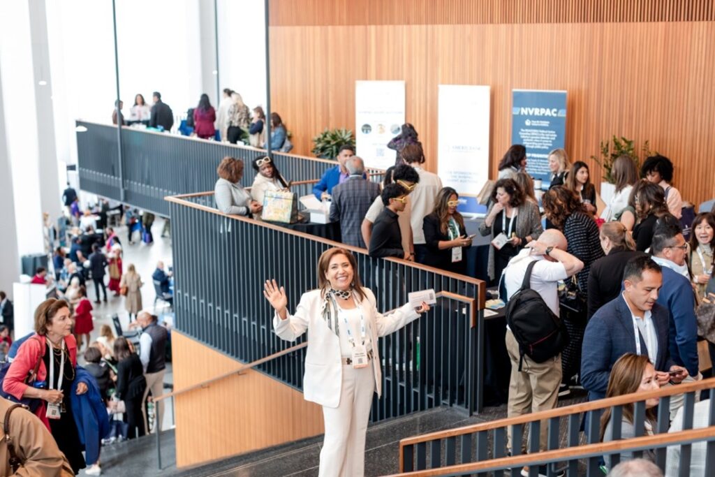 People gather at a modern conference venue, networking and engaging in discussions. A person in front smiles and waves, holding papers, creating a lively atmosphere.