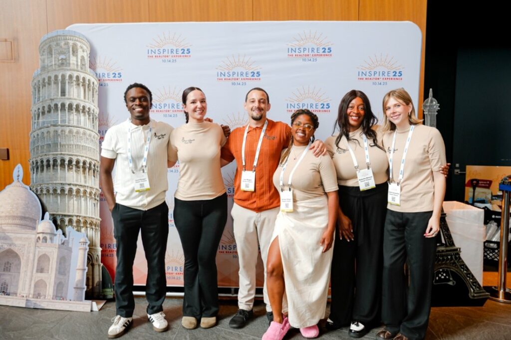 A group of six people stands smiling in front of a banner with Pisa, Taj Mahal, and Eiffel Tower images at an event.