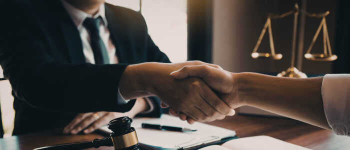 Two people shaking hands across a wooden desk with legal documents, gavel, and scales of justice visible, suggesting a professional legal setting.
