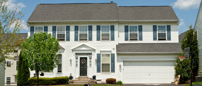 A two-story suburban house with white siding, black shutters, and a double garage, surrounded by green trees and a clear blue sky.
